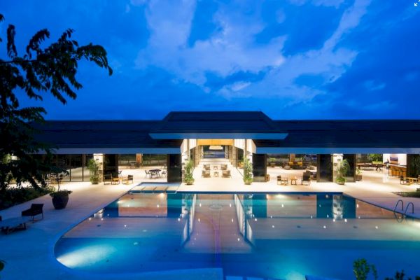 A modern, well-lit building with a large swimming pool in front, set against a beautiful evening sky, surrounded by greenery and illuminated pathways.