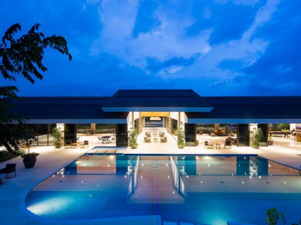 A modern, well-lit building with a large swimming pool in front, set against a beautiful evening sky, surrounded by greenery and illuminated pathways.