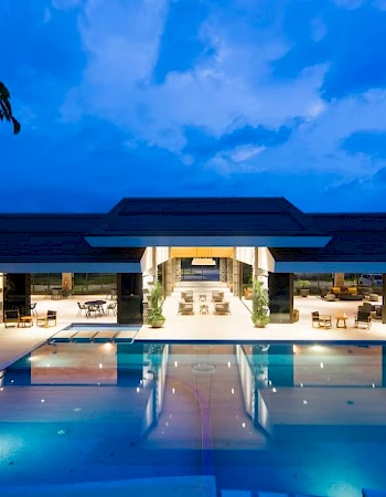 An outdoor pool area is illuminated at night, leading to a modern, open-concept building with seating areas and large glass windows.