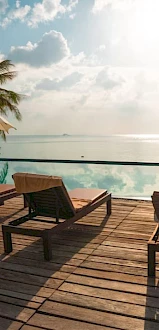 A serene poolside view with lounge chairs, umbrellas, palm trees, and a tranquil sea under a partly cloudy sky, portraying a relaxing atmosphere.