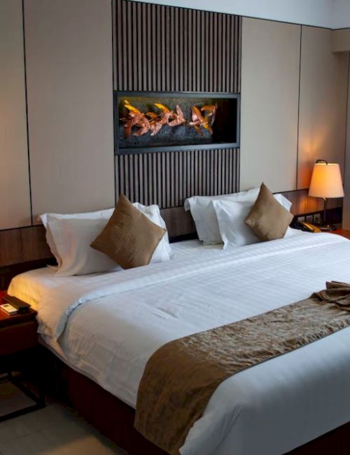 A well-lit hotel room featuring a neatly made king-sized bed with decorative pillows, bedside lamps, and a small table beside the bed.