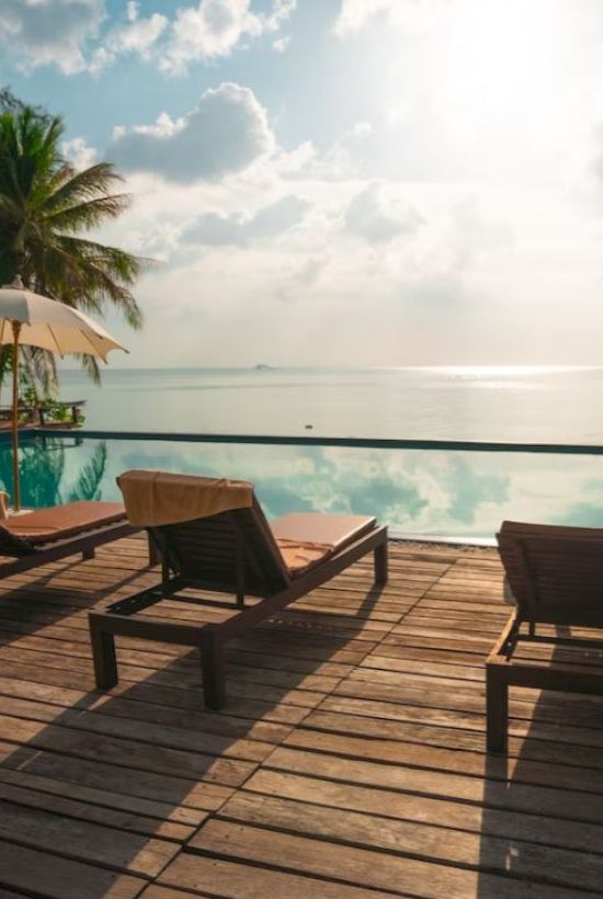 A serene poolside scene with lounge chairs, umbrellas, and palm trees overlooking a calm sea under a partly cloudy sky.