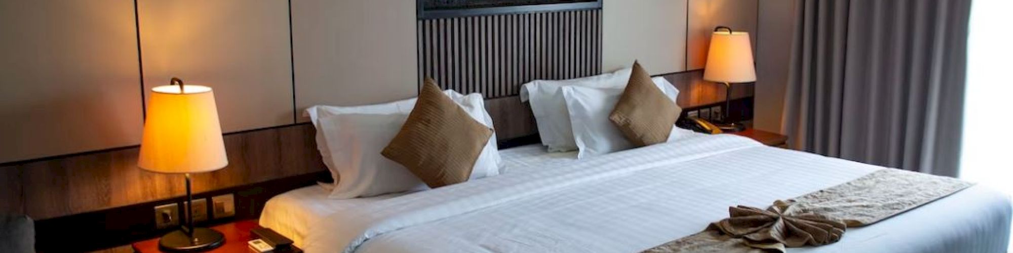 A neatly arranged hotel room featuring a large bed with pillows and lamps on both sides, a wall art piece, and a window with closed curtains.