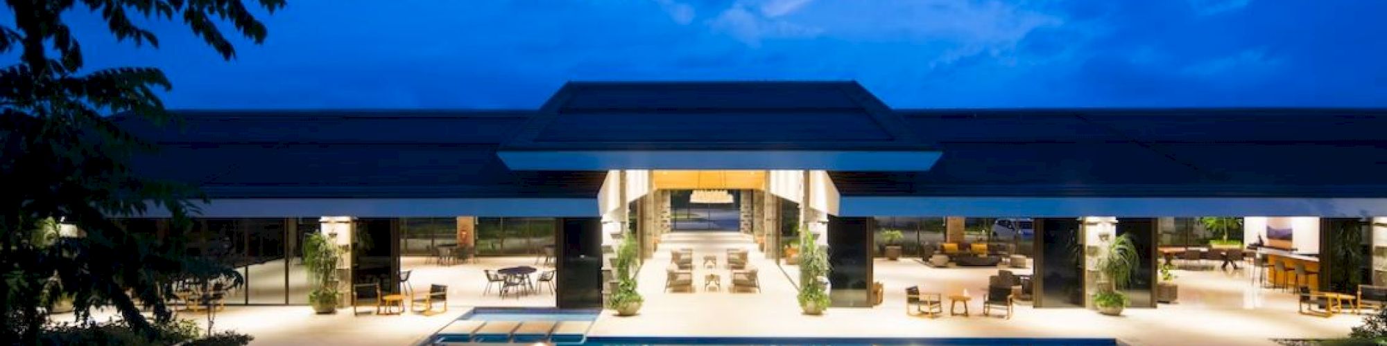 The image shows a modern building with an illuminated swimming pool in the foreground, set against a twilight sky.