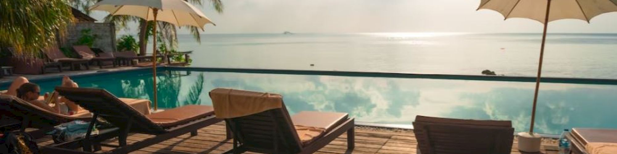 The image shows a serene poolside scene with lounge chairs, umbrellas, and a view of the ocean and sky, offering a tropical getaway atmosphere.