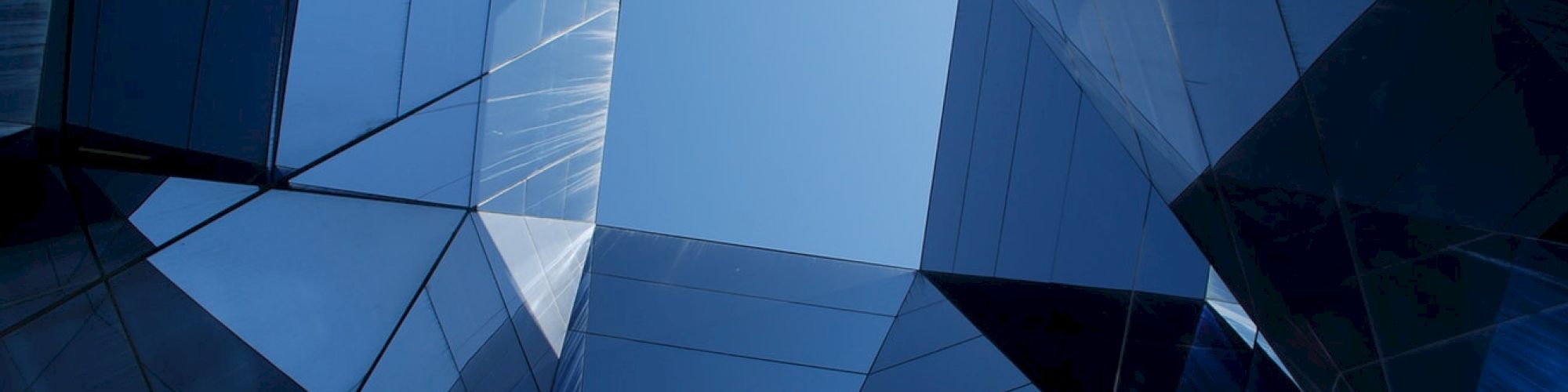 The image showcases an upward view of modern angular glass buildings reflecting the sky, creating a geometric pattern with sharp lines and edges.
