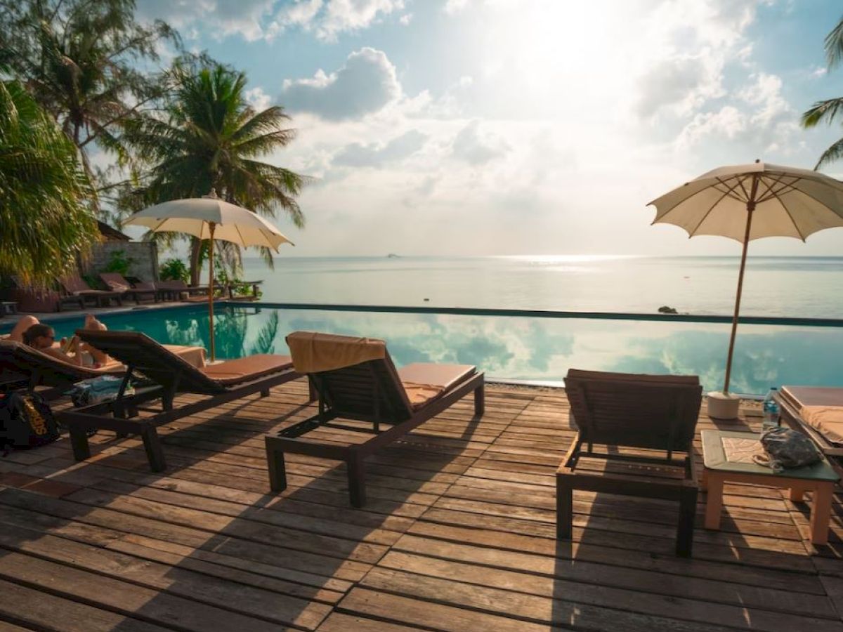 A serene poolside scene with lounge chairs, parasols, and palm trees, overlooking a calm body of water with a bright, partly cloudy sky.