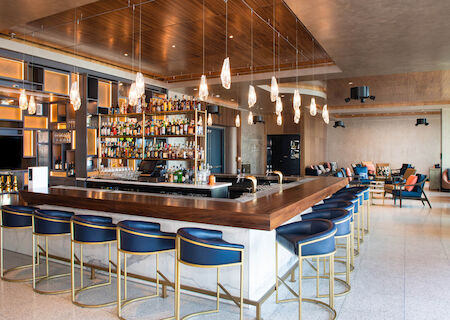 This image shows a modern bar with blue barstools, a well-stocked liquor shelf, and a cozy lounge area with seating in the background.
