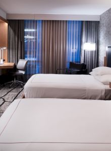 A modern hotel room features two double beds, a flat-screen TV, a desk with a chair, and a window with drapes and a view, all under ambient lighting.