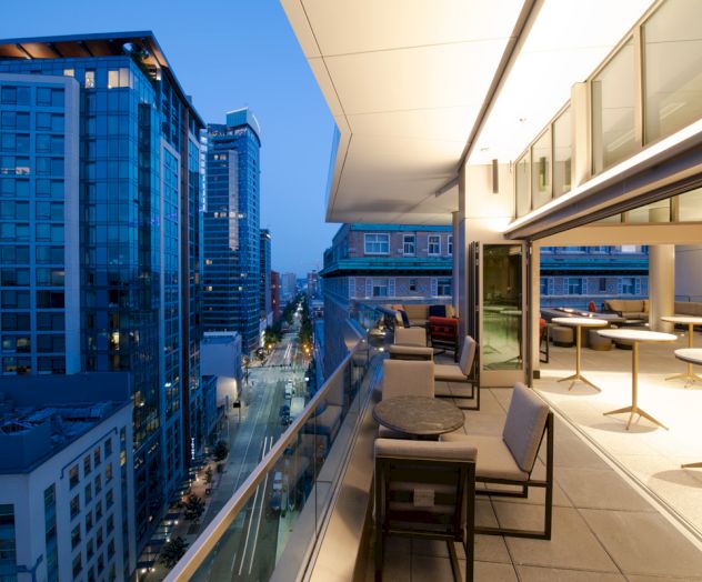 The image shows a modern balcony with seating and tables overlooking a cityscape with tall buildings and a street below at dusk.