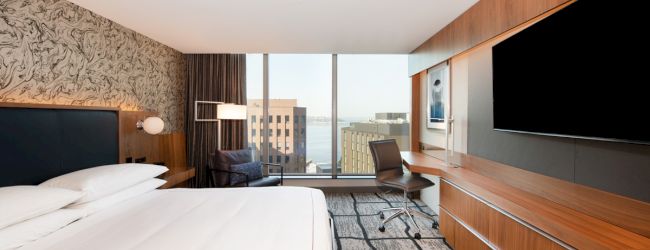 A modern hotel room with a large bed, wooden furnishings, a desk, a chair, and a large wall-mounted TV, with windows showing a cityscape view.