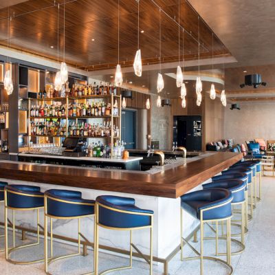 A modern bar with wooden accents, sleek lighting, blue bar stools, and a fully stocked liquor shelf; an inviting and stylish setting.