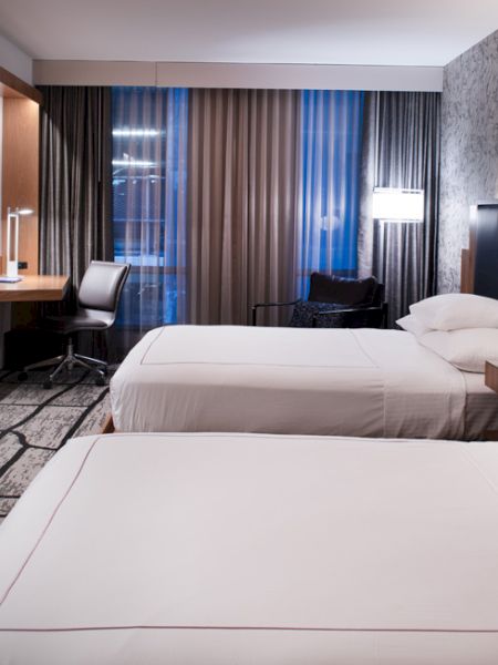 A modern hotel room with two beds, a flat-screen TV, desk, chairs, and bedside lamps, with a patterned carpet and wallpaper to match.