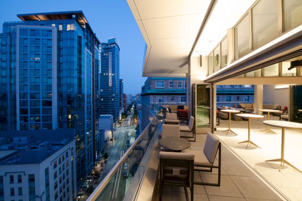 A modern balcony with furniture overlooks a city street lined with tall buildings during twilight, showcasing a mix of city lights and the evening sky.