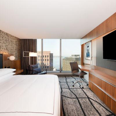 A modern hotel room with a large bed, a desk, a TV, and a view of city buildings through large windows, featuring contemporary decor.
