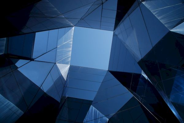 The image shows a geometric scene with reflective glass surfaces forming triangular patterns, allowing a glimpse of blue sky through the center.