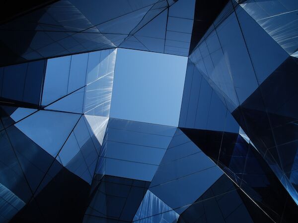 The image shows a geometric scene with reflective glass surfaces forming triangular patterns, allowing a glimpse of blue sky through the center.
