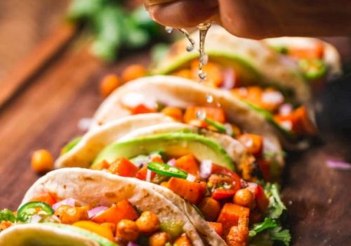 A hand squeezes fresh lime juice over a row of vibrant veggie tacos filled with chickpeas, diced tomatoes, cilantro, and avocado, all arranged on a wooden board.