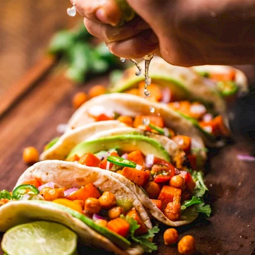 A hand squeezes fresh lime juice over a row of vibrant veggie tacos filled with chickpeas, diced tomatoes, cilantro, and avocado, all arranged on a wooden board.