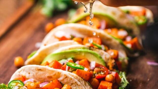 A hand squeezes fresh lime juice over a row of vibrant veggie tacos filled with chickpeas, diced tomatoes, cilantro, and avocado, all arranged on a wooden board.