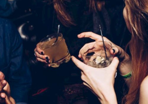 People are socializing and holding drinks, with one person laughing and others reaching towards them. Only their upper bodies and hands are visible.