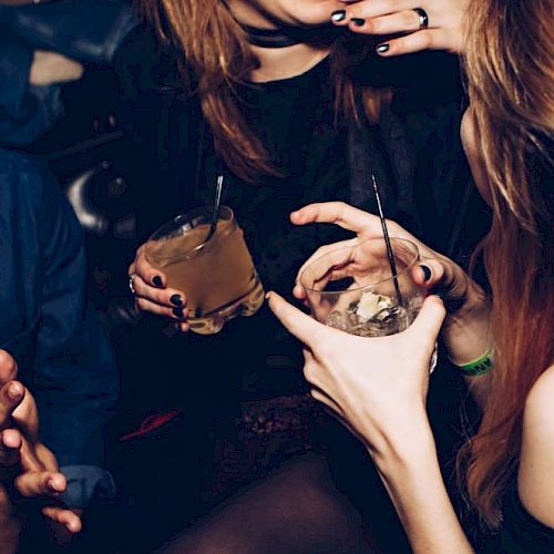 People are socializing and holding drinks, with one person laughing and others reaching towards them. Only their upper bodies and hands are visible.
