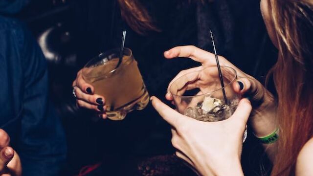 People are socializing and holding drinks, with one person laughing and others reaching towards them. Only their upper bodies and hands are visible.