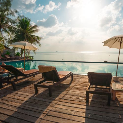 Poolside with lounge chairs, umbrellas, and a wooden deck overlooking a serene sea at sunset, surrounded by tropical trees and clear skies.