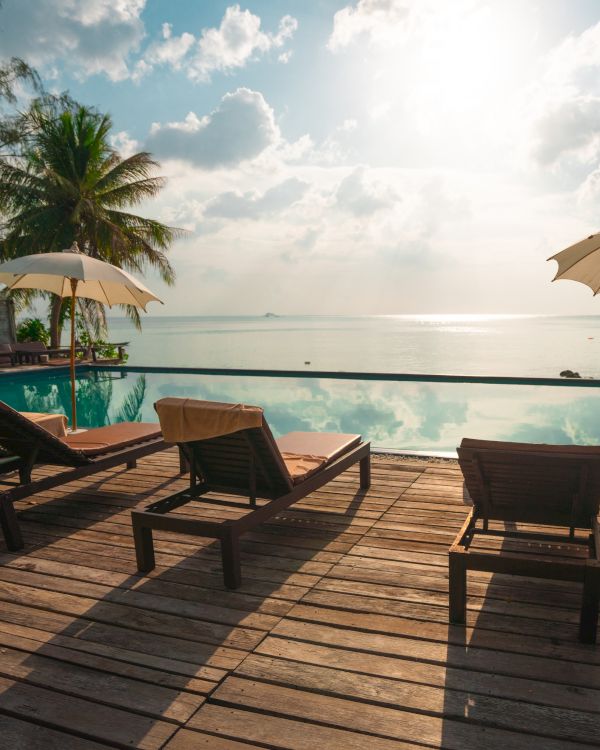 Poolside with lounge chairs, umbrellas, and a wooden deck overlooking a serene sea at sunset, surrounded by tropical trees and clear skies.