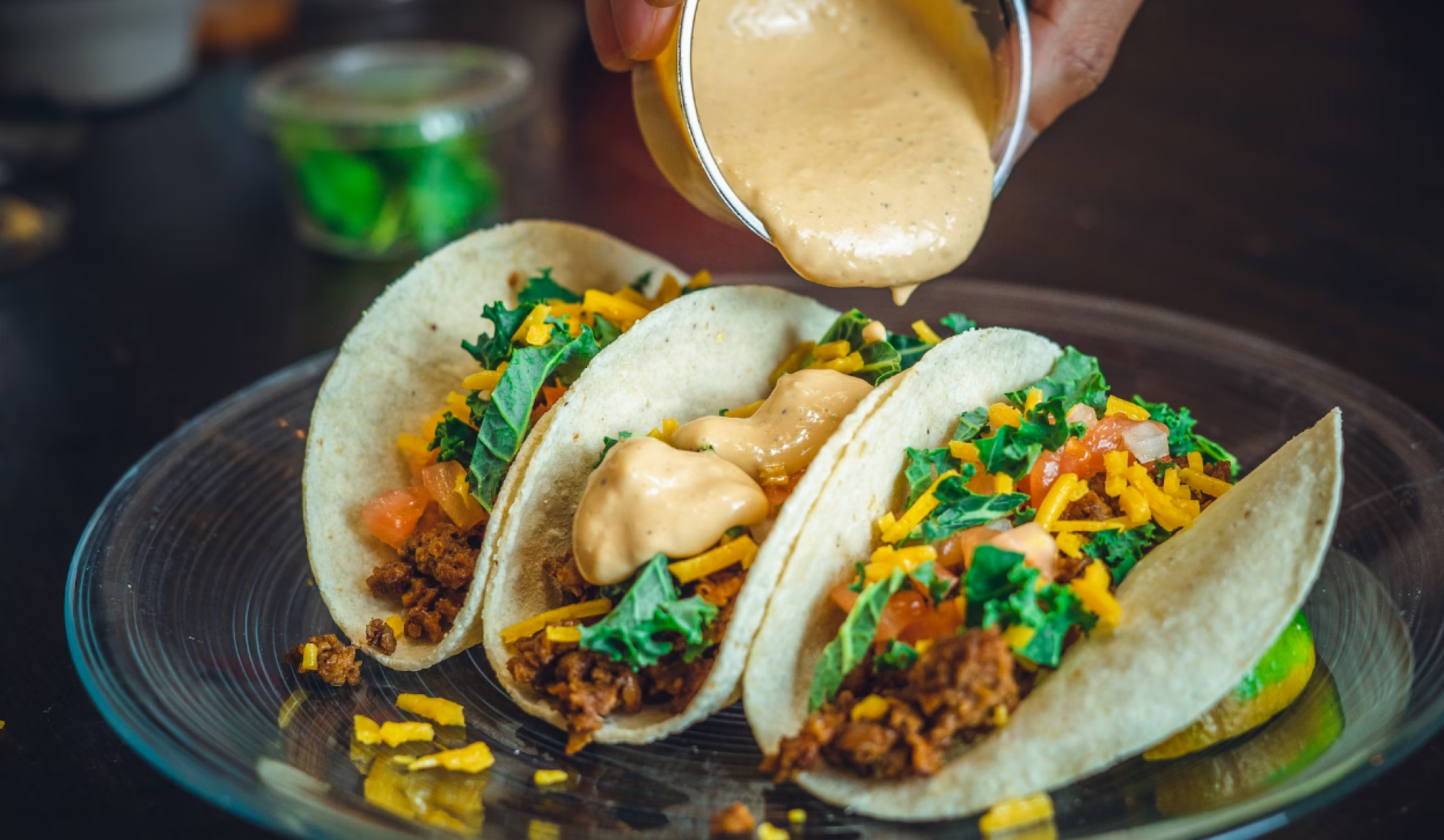 A hand is pouring a creamy sauce over three tacos on a plate, filled with meat, shredded cheese, greens, and diced tomatoes.