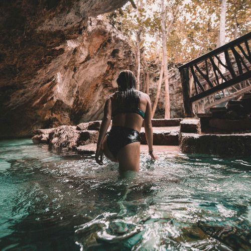 A person in swimwear walks through a clear water cave, with steps and lush greenery visible in the background under soft, warm lighting.