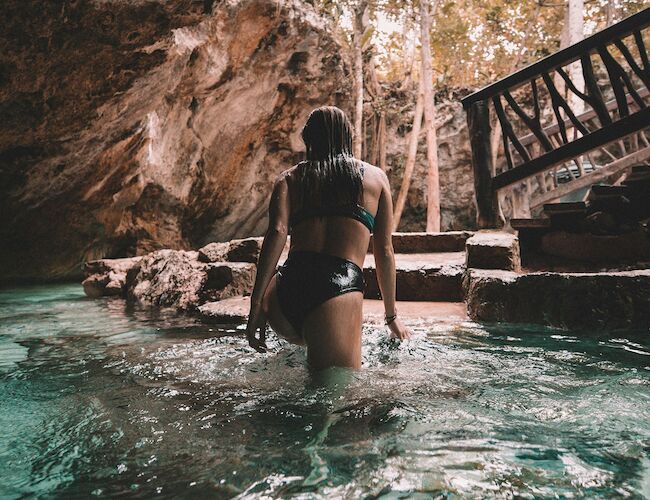 A person in swimwear walks through a clear water cave, with steps and lush greenery visible in the background under soft, warm lighting.