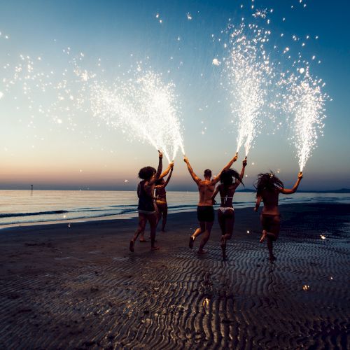 People on a beach at dusk holding sparklers with firework-like sparks, creating a festive atmosphere by the shoreline, possibly celebrating.