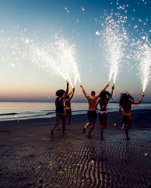 People on a beach at dusk holding sparklers with firework-like sparks, creating a festive atmosphere by the shoreline, possibly celebrating.