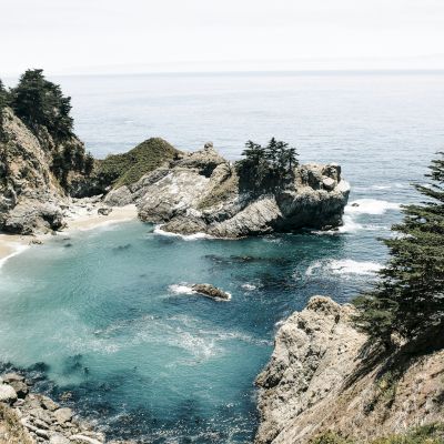 The image shows a scenic coastal landscape featuring a small, sandy beach, clear blue water, rocky cliffs, and trees on a sunny day.