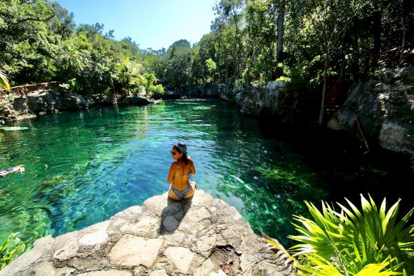 A person sits on a rock ledge overlooking a clear blue-green lagoon surrounded by lush greenery and tall trees under a bright sky.