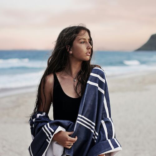 A person stands on the beach during sunset, wearing a black outfit and wrapped in a blue and white striped towel, looking off into the distance.