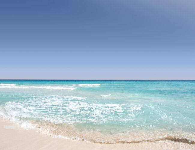 The image shows a serene beach with clear blue skies and turquoise water gently washing onto the sandy shore.