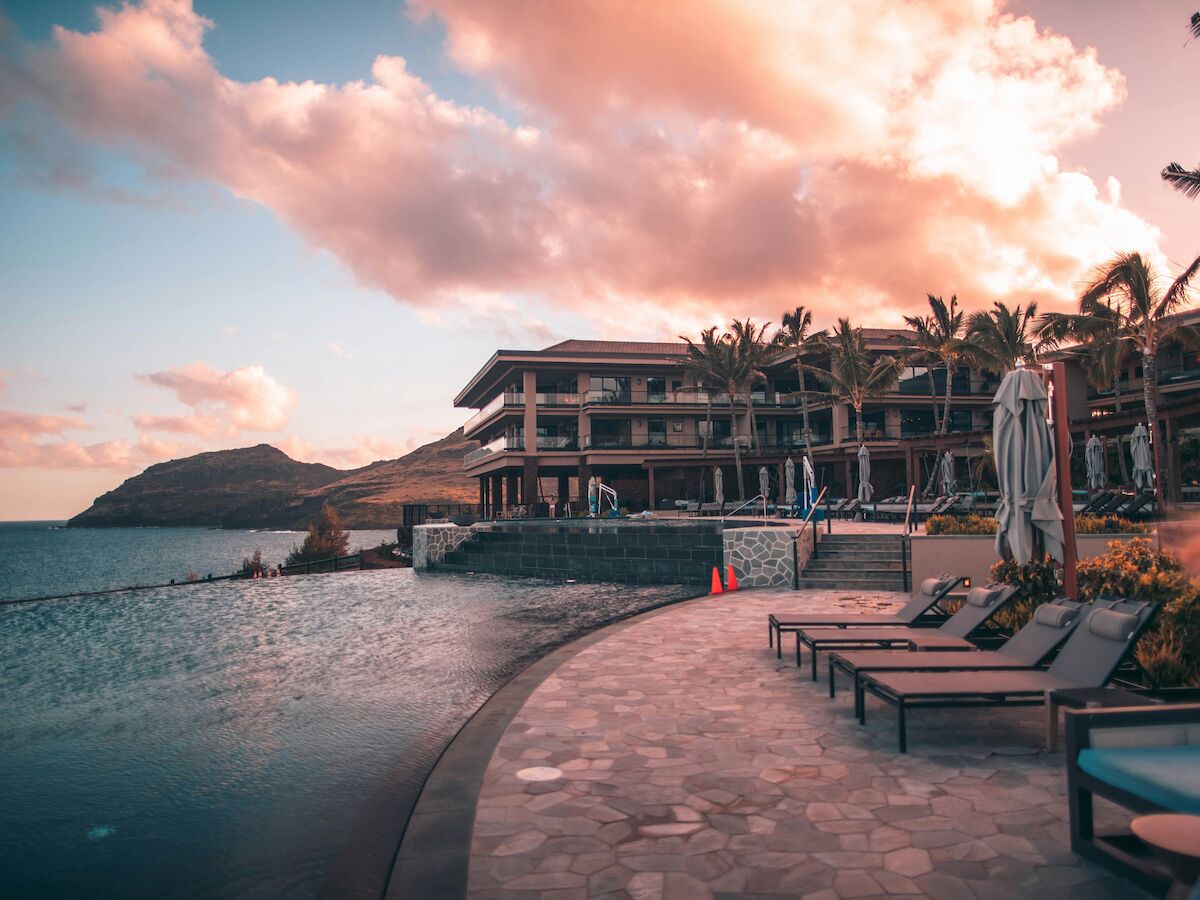 The image shows a coastal resort with a pool, lounge chairs, and palm trees under a vibrant sky during sunset.