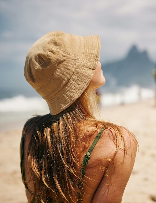 A person with long hair in a sun hat sits on a beach, facing the ocean. The sky is cloudy, and it's a serene beach scene.