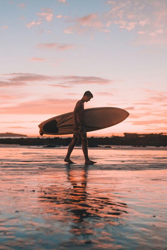 A person walks along the beach with a surfboard during a colorful sunset, reflected in the wet sand and creating a serene scene.