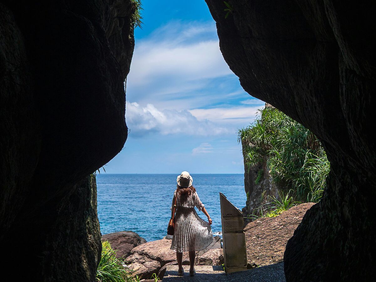 A person stands at the entrance of a cave, looking out at the ocean under a bright blue sky with fluffy clouds.