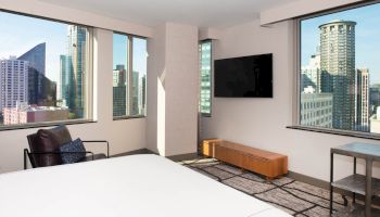 This image shows a modern hotel room with large windows, offering a city skyline view, a television, a chair, a low wooden table, and a bed.