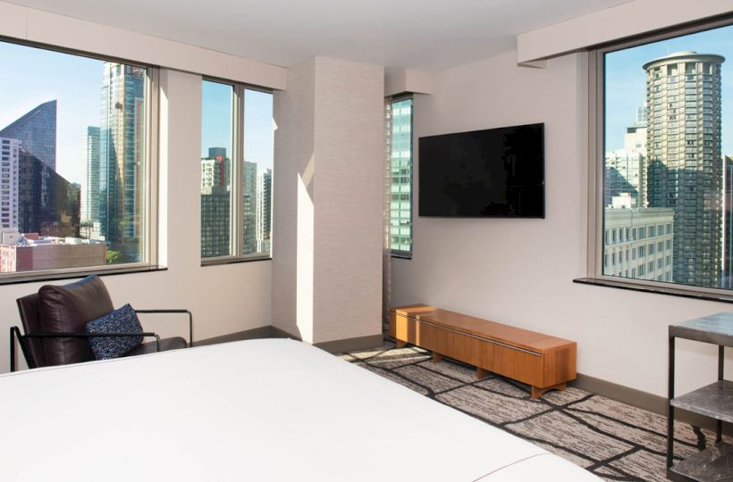 This image shows a modern hotel room with large windows, offering a city skyline view, a television, a chair, a low wooden table, and a bed.