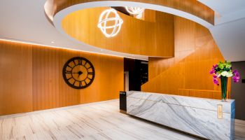 A reception area with a marble desk, wooden walls, a large wall clock, modern lighting fixtures, and a flower arrangement on the desk.