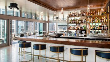 This image shows a modern bar area with high stools, a wooden counter, and shelves stocked with various bottles of alcohol, overlooking a city view.