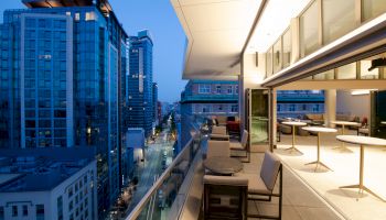 A view from a balcony of a modern high-rise building, overlooking a city street lined with other tall buildings at dusk, with outdoor seating.