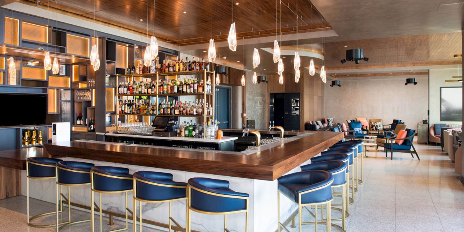 A modern bar with a wooden counter, blue stools, a display of various bottles, hanging lights, and a seating area in the background.