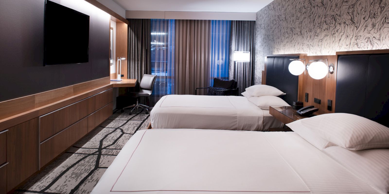 A modern hotel room featuring two neatly made beds, a flat-screen TV, a work desk with a chair, and contemporary decor with accent lighting.