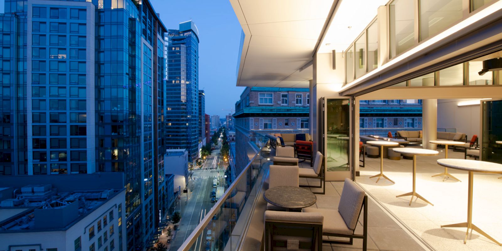 The image shows a modern balcony with seating, overlooking a city street lined with high-rise buildings at dusk, with interior dining tables visible.